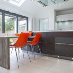 Functional wood effect laminate breakfast table seating on kitchen island assesorised with orange dining chairs.