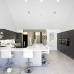 A grand open plan living area bosting a high end modern kitchen in netural light grey and graphite with white worktops.